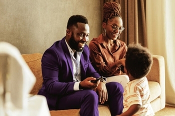 Three people gather in a comfortable living room with warm natural light streaming through a large window. Two adults sit on a sofa, facing a young child who stands nearby. 